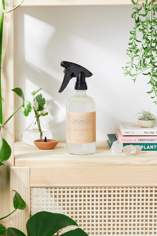 Glass spray bottle with a label on a wooden shelf surrounded by plants and decor
