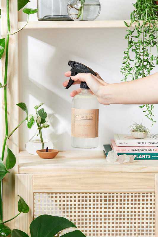 Person holding a 'Greenics' spray bottle on a shelf with plants and books.