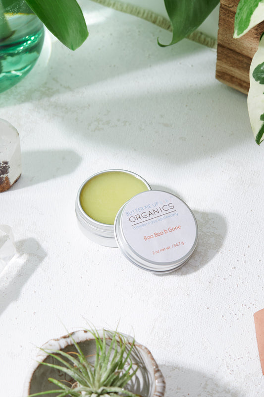 Small container of green cream with a white lid on a light surface, surrounded by plants.