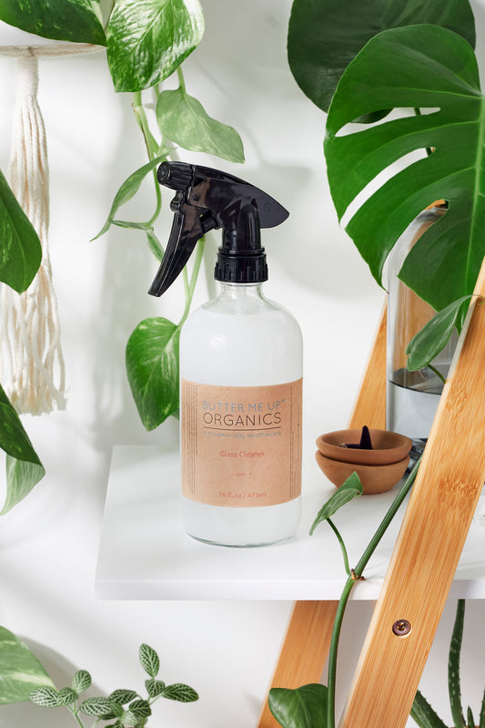Clear spray bottle labeled 'Organics' with a brown label on a white surface surrounded by green plants.