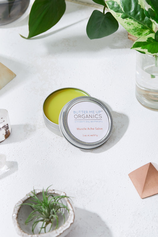 Butter Me Up Organics salve tin on a white surface with plants in the background
