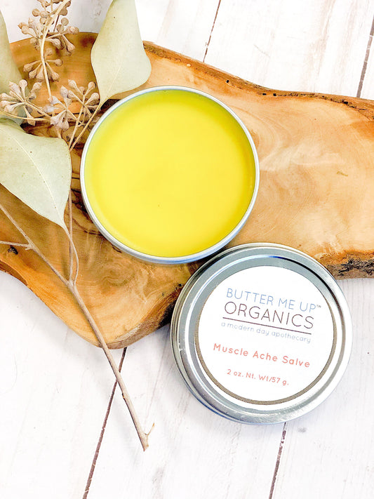 Two tins of Butter Me Up Organics Muscle Ache Salve on a wooden board with leaves.