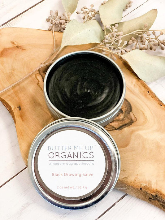 Butter Me Up Organics Black Drawing Salve in a metal tin on a wooden surface with dried leaves.