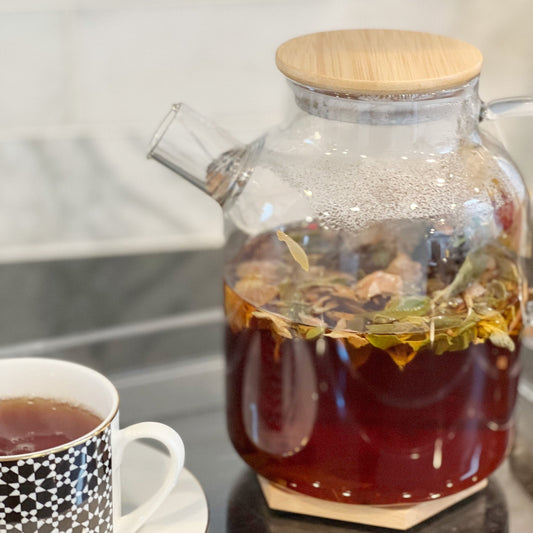 Glass teapot with wooden lid containing a hot beverage, next to a cup of tea.