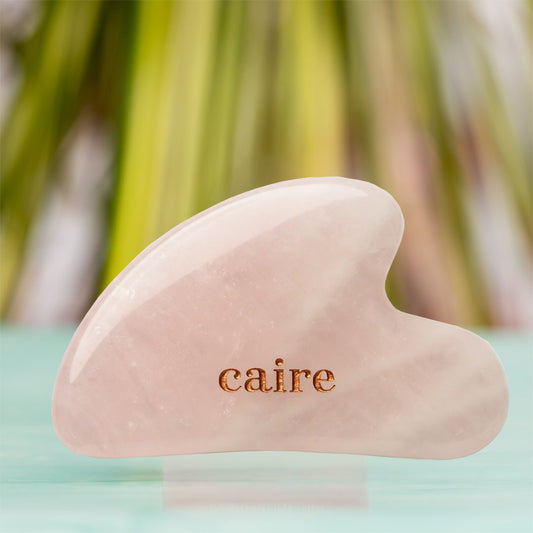 Pink gua sha tool with 'caire' branding on a blurred natural background