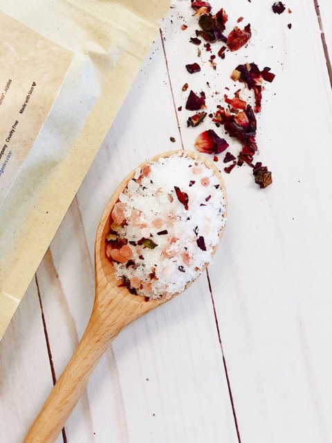 Wooden spoon with bath salts and dried herbs on a light wooden surface