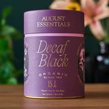 Purple canister of August Essentials Decaf Black Organic Black Tea on a wooden surface with a blurred floral background.