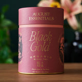 August Essentials Black Gold Organic Black Tea canister on a wooden surface with a blurred floral background