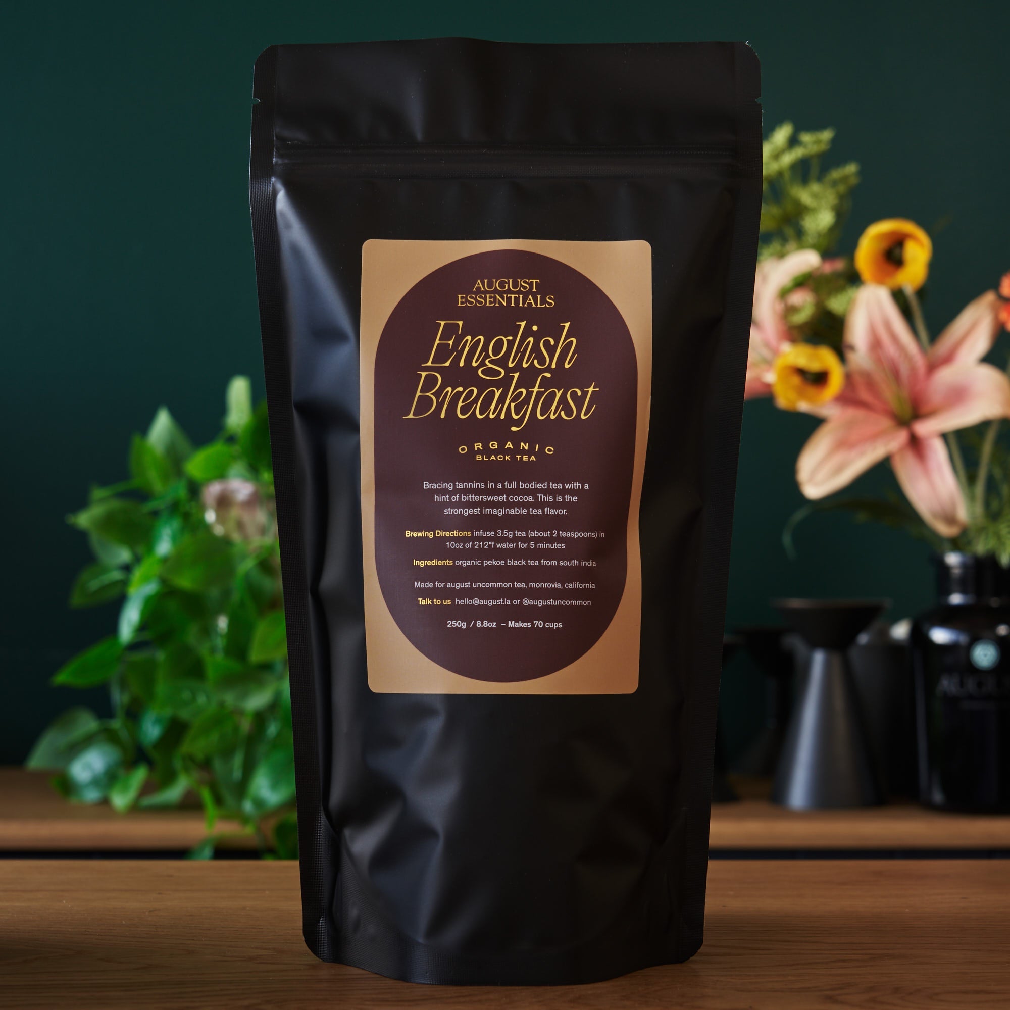 Black tea package labeled 'English Breakfast' by August Essentials on a wooden surface with plants in the background.