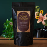 Black tea package labeled 'English Breakfast' by August Essentials on a wooden surface with plants in the background.