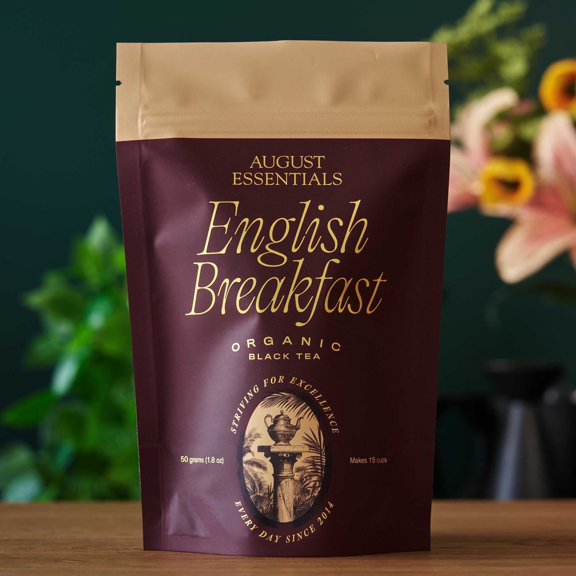 August Essentials English Breakfast organic black tea package on a wooden surface with a blurred floral background