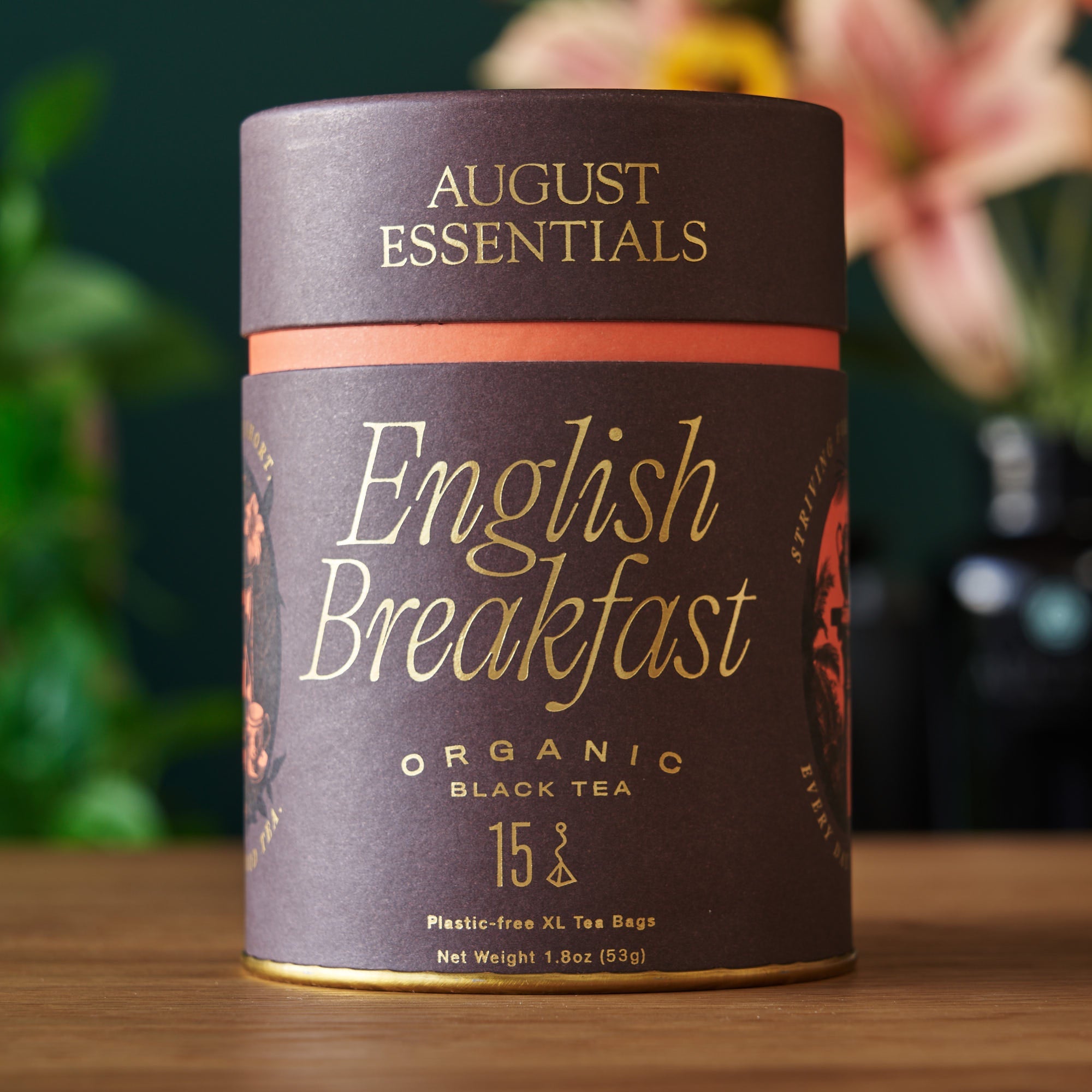 August Essentials English Breakfast organic black tea canister on a wooden surface with a blurred floral background