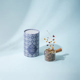Candle with decorative packaging and a small container of dried herbs on a light blue background