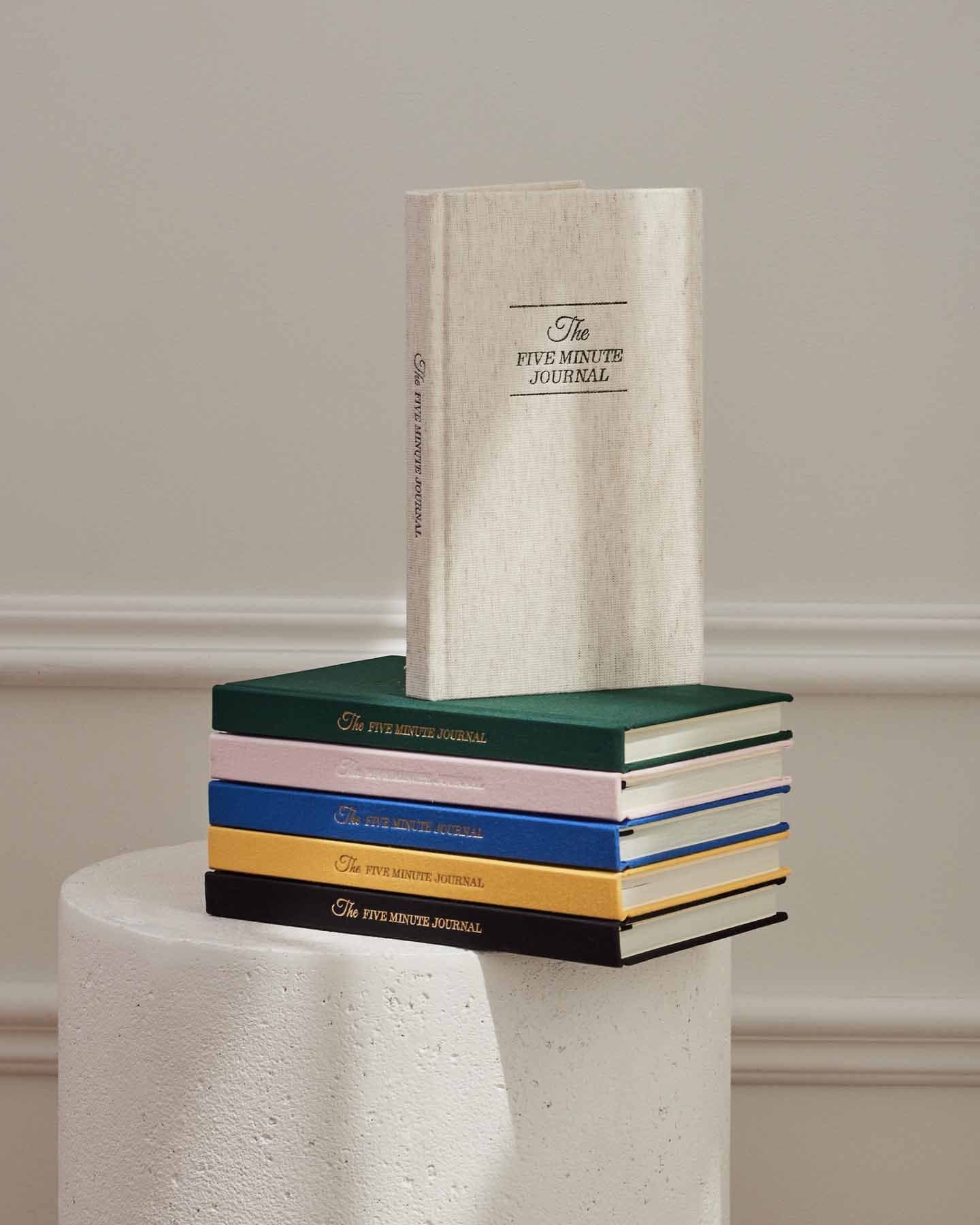 Stack of books titled 'The Five Minute Journal' on a pedestal against a neutral background