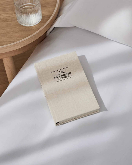 Beige notebook titled 'The Five Minute Journal' on a white surface with a wooden side table in the background.