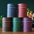 Five August Essentials tea canisters on a wooden surface with a blurred floral background.