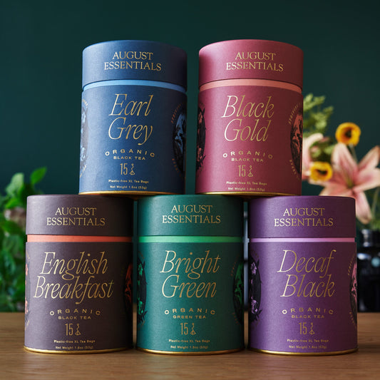 Five August Essentials tea canisters on a wooden surface with a blurred floral background.