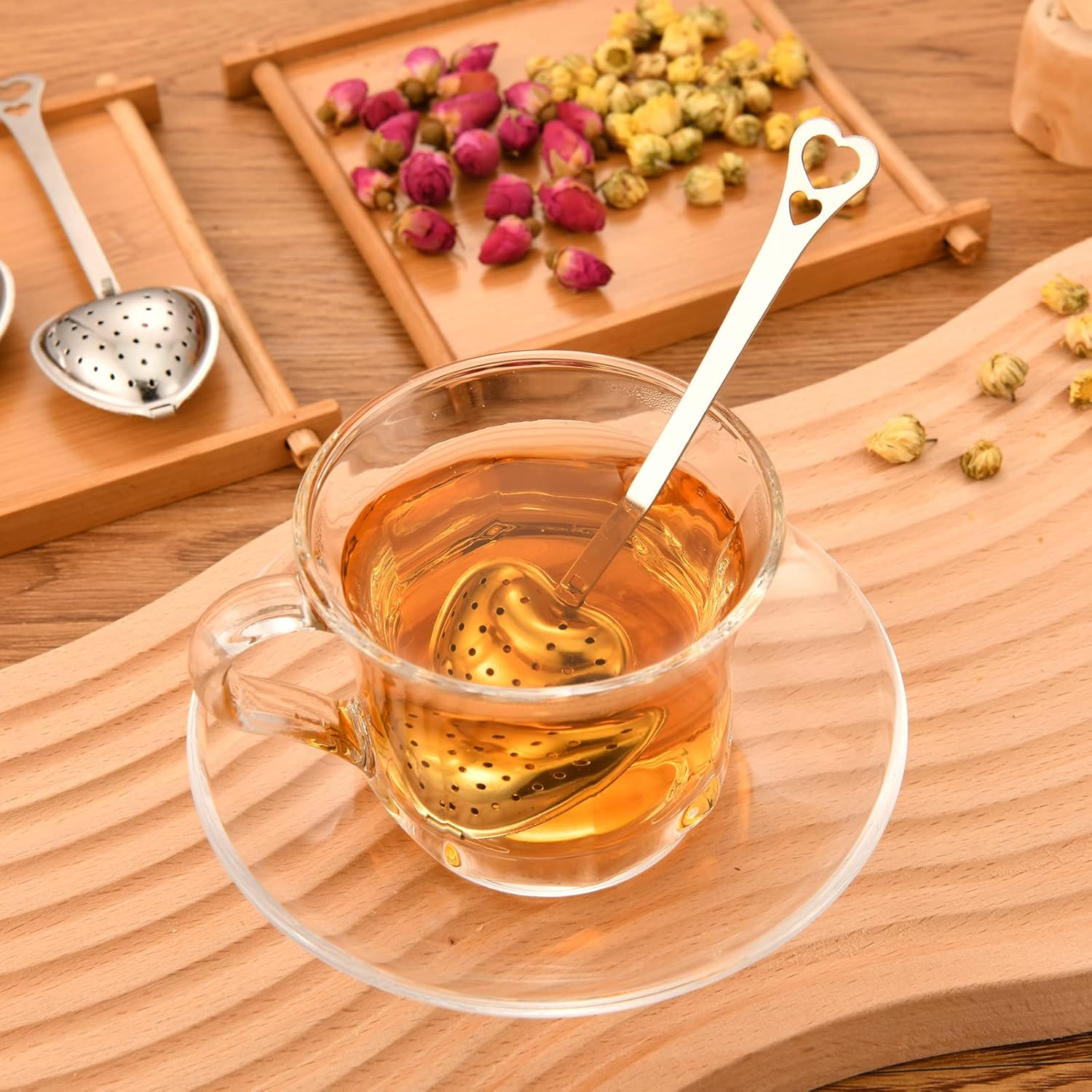 Clear glass cup of tea with a heart-shaped tea infuser on a wooden surface with dried tea leaves.