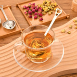 Clear glass cup of tea with a heart-shaped tea infuser on a wooden surface with dried tea leaves.