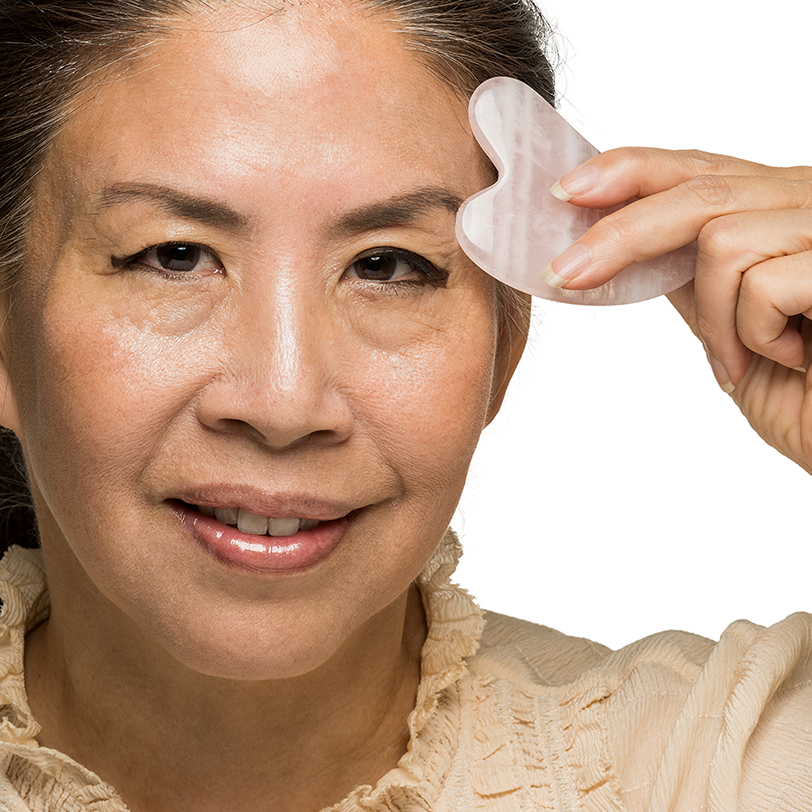Woman using a pink gua sha tool on her face against a white background