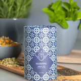 Blue and white patterned tea canister labeled 'Sleeping Herbal Tea' on a wooden surface with plants in the background.