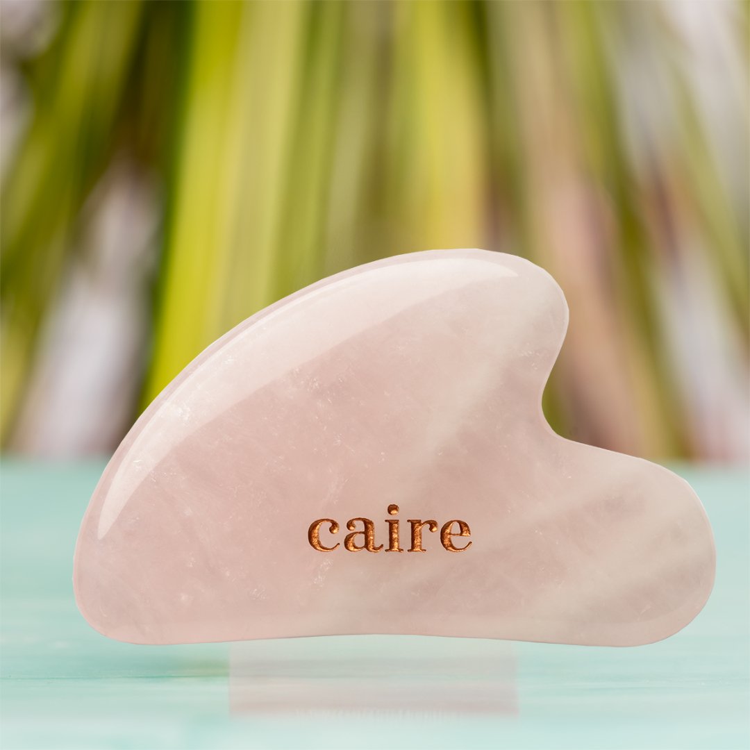 Pink gua sha tool with 'caire' branding on a blurred natural background